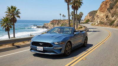 A blue Ford Mustang Convertible driving along the sunny Pacific Coast Highway with the ocean and cliffs in the background.