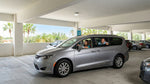 A silver Chrysler Pacifica minivan is being parked by a woman in a parking garage, with a theme park archway and palm trees visible in the background.