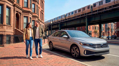 Couple walking in Boston near a silver car rental and elevated train, comparing driving vs public transit.