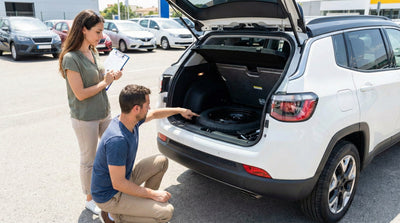 A man kneeling and checking the spare tyre compartment under the trunk floor of a white Toyota Rav4 SUV in a car rental lot.