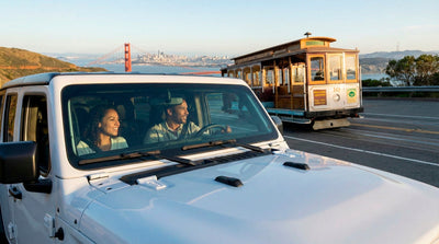 Couple driving a white Jeep rental car in San Francisco passing a cable car with the Golden Gate Bridge behind.