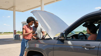 A couple at a rural Texas gas station is checking the engine of a grey Jeep Grand Cherokee rental car, with a warning light visible on the dashboard.