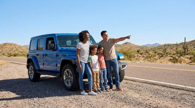 Un grupo familiar disfruta de una parada escénica soleada junto a su auto de alquiler azul, con vista a un paisaje de carretera abierta.